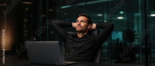 Content businessman relaxing at his desk in a modern dark office at night after work.
