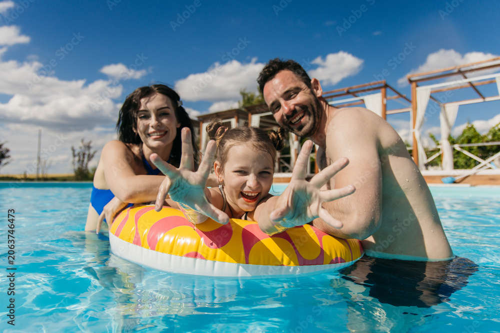 Obraz premium Young family enjoying time together in swimming pool, smiling and playing with inflatable ring under clear blue sky and bright sunlight