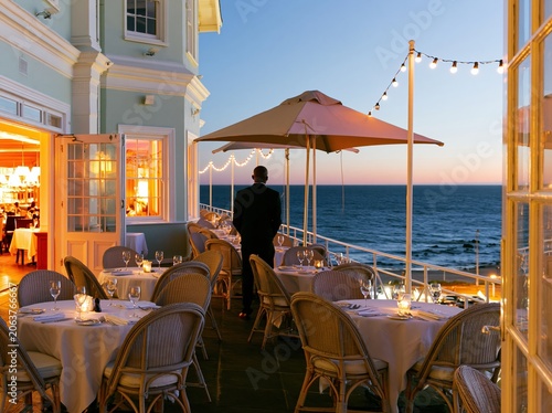 Luxury coastal restaurant terrace at sunset with a person looking at the sea for travel promotion, fine dining advertising and vacation hospitality background