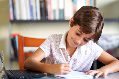 Disciplined schoolboy attentively works on his homework with laptop and reading materials nearby