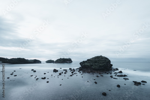 Photography Long exposure seascape of rocky formations and scattered boulders in calm, misty water under an overcast cloudy sky