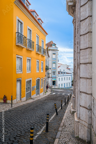 Historic Alfama street in Lisbon, Portugal, showing a vibrant yellow building, traditional Portuguese pavement, and classic architecture under a bright blue sky
