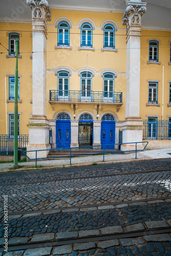 Lisbon street featuring a historical yellow building facade with blue doors and windows, intricate columns, a balcony, and traditional cobblestone pavement with tram tracks