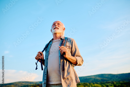 Happy senior man with a backpack enjoying an outdoor adventure, looking up at the clear blue sky, symbolizing freedom and vitality