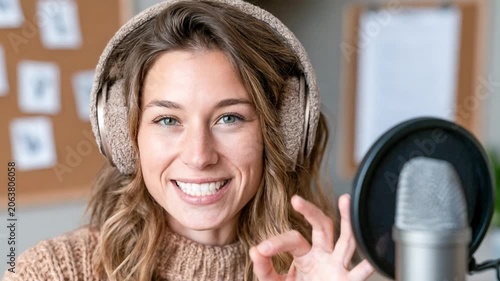 Woman Wearing Headphones by Microphone: A young woman with headphones on and smiling, making an okay sign with her fingers, sits in front of a microphone.