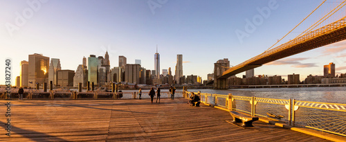 People walking and sitting on a wooden pier viewing the skyline at sunset, representing an urban travel lifestyle by the East River, Lower Manhattan, Brooklyn Bridge, New York City.
