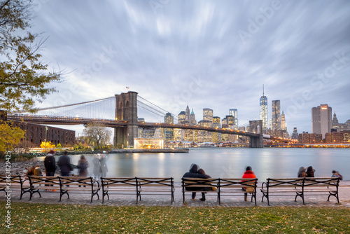 Travelers and residents relax on park benches at dusk, capturing the scenic urban lifestyle and city exploration in New York City, East River, Lower Manhattan, Brooklyn Bridge.