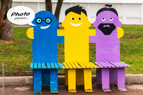 Colorful wooden bench shaped like three hugging friends in a park. Creative public sitting spot with photo please speech bubble. Outdoor seats stylized human characters with smiling faces.