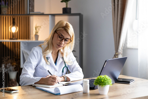 Quadro su tela Female doctor in white coat and stethoscope writing medical notes in a notebook