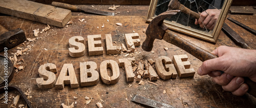 Hand striking wooden dimensional letters spelling self sabotage with a claw hammer, creating wood chips on a rustic woodworking bench