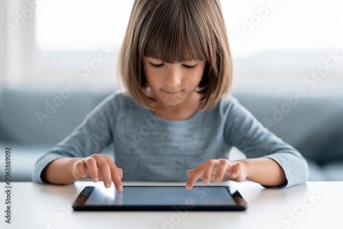 Young girl with short brown hair interacts with a tablet device on a white table, seated in a bright room with soft furnishings and natural light filtering through the window in the background
