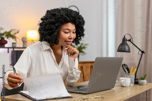 Canvas Print African American businesswoman at home office matches documents to laptop data smiling satisfied