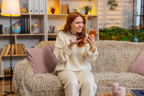 Vászonkép Young woman on home sofa talks on smartphone speaker and waves hand in furious dispute showing frustration