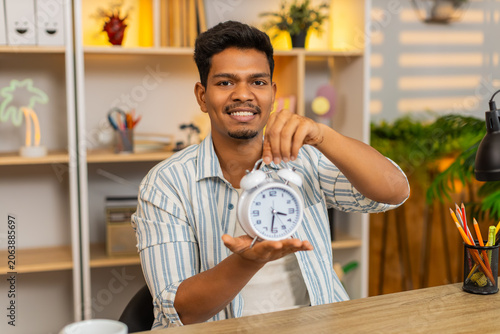 Indian man on home table shows clock to camera and talks with tense face about time during remote work task. Arabian guy on home table explains delay and asks about schedule in calm home office mood.