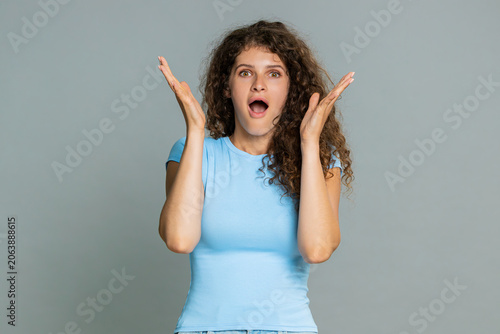 Quadro su tela Young woman looking surprised, opening mouth widely and touching cheeks expressing astonished reaction to unexpected event