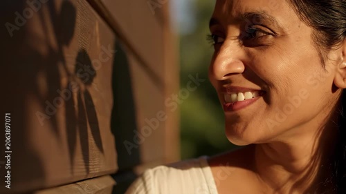 Smiling Indian woman standing outdoors against wooden wall. Close up portrait of happy lady in sunshine with leaf shadow projection on siding. Lifestyle concept