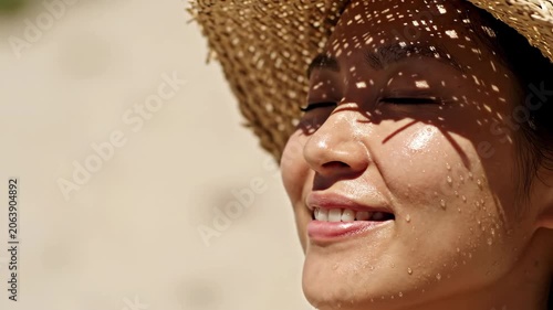 Asian woman wearing a straw hat on a beach. Female model with sun-kissed skin and water droplets on her face relaxing under sunlight. Summer travel skin care