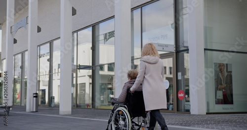 Caregiver pushing wheelchair with elderly person outside modern building. Care and assistance concept. Daytime scene with urban environment.