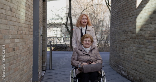 Young caregiver assisting elderly woman in wheelchair outdoors on sunny day. Providing support and companionship during stroll. Concept of caregiving, mobility assistance, and senior care.