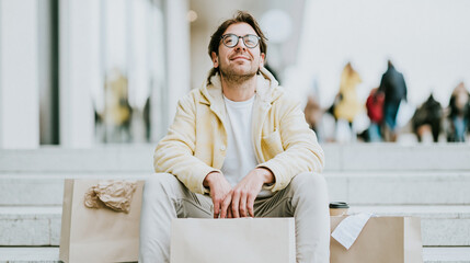 Young man sitting on store steps with plain shopping bags no logos relaxed posture coffee cup nearby wrinkled receipt on one bag street background and passersby defocused