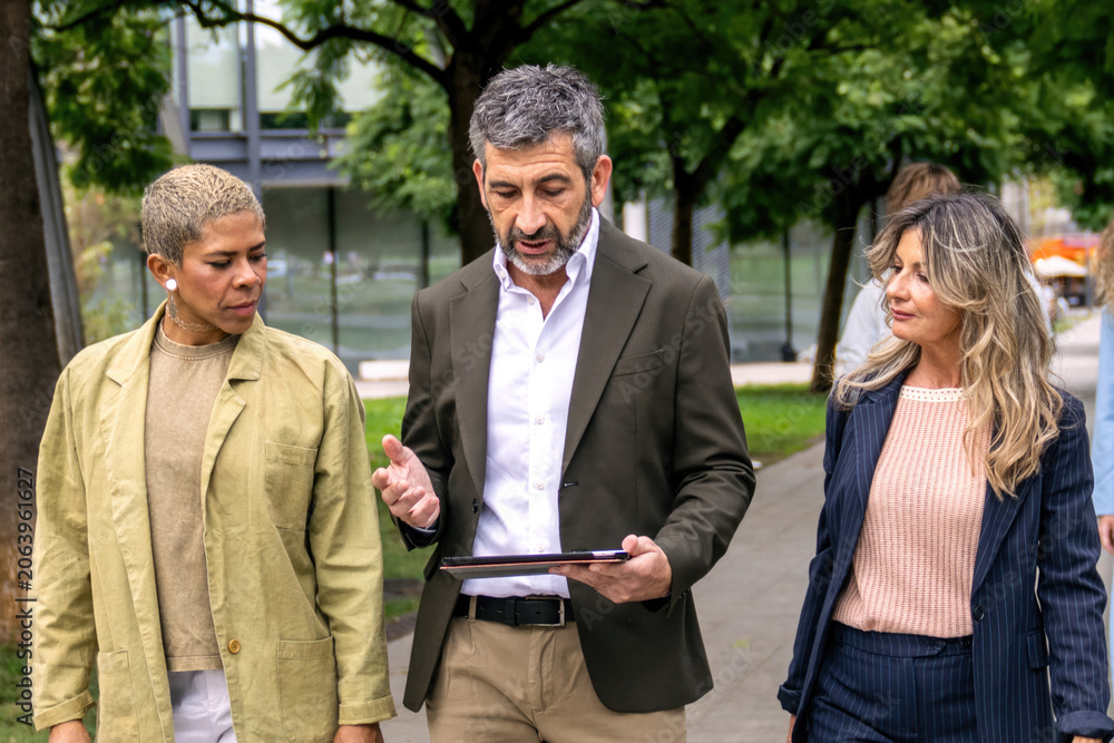 Obraz premium Group of diverse business people walking outdoors, having a discussion and looking at a tablet during a casual meeting