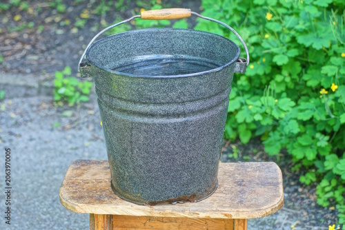 one black metal bucket with water stands on a brown wooden table on a summer street