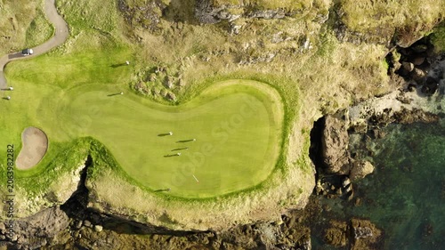 An amazing top-down aerial view of golfers playing on a beautiful green course situated on a rocky cliffside next to the clear ocean water.