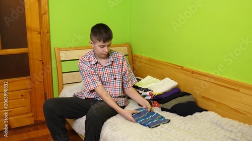 A boy sits on the bed folding clothes, organizing his room and neatly arranging items around him.