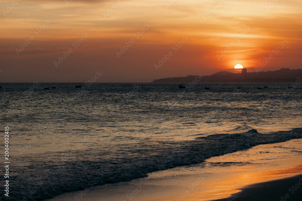 Obraz premium Sun sets over a tranquil ocean with distant hills silhouetted against an orange sky. Mui Ne, Vietnam
