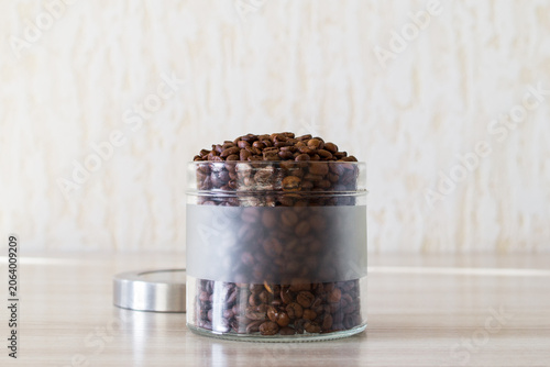 Open glass jar with coffee beans on wooden table in light kitchen. Preparing for brewing coffee.