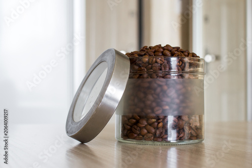 Glass jar of freshly roasted coffee beans on wooden table with natural light at home interior.