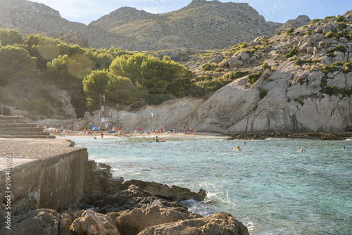 Εκτύπωση καμβά Rocky beach with turquoise waters and sunbathers beneath lush green hills