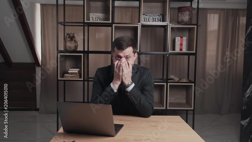 Tired man in a black suit with a laptop on the table covers his face with his hands and rubs his eyes. Night broadcast. Sleepy presenter of the educational channel.