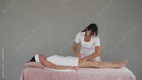 Professional masseuse in white uniform performs relaxing leg massage on client lying on pink table in spa salon. Therapist applies oil and strokes legs with hands