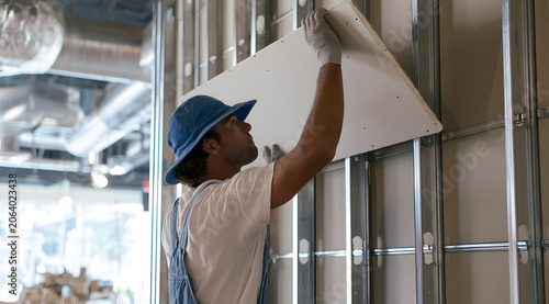 Drywall worker affixing gypsum panels to metal framing on a wall, showcasing drywall installation, an interior finish, and commercial building work