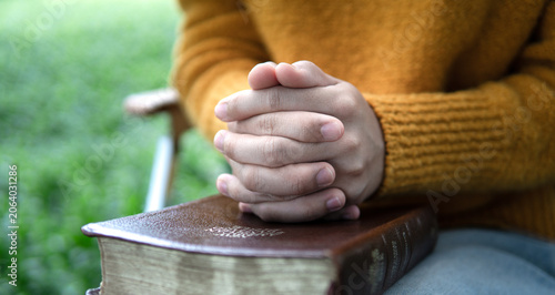 Fotografie A close-up image of hands clasped together in a prayerful posture, looking at a Bible outdoors against a green background