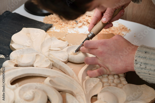 A close-up of a craftsman hands using chisel to carve an ornate wooden panel, surrounded by wood shavings. Detailed woodworking scene emphasizing traditional craft, skillful hand carving