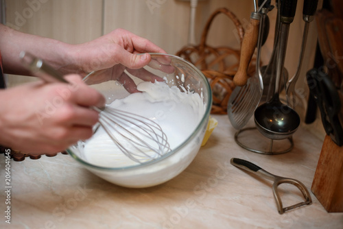 Hands whisking cream in a glass bowl on kitchen counter, surrounded by utensils and a peeler, capturing the homemade baking process. Son helps mom at home, whips cream for pastries