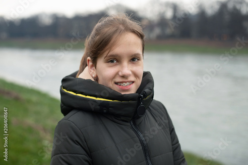 A smiling girl with braces stands beside a river on overcast day wearing a padded winter coat. The girl is embarrassed to wear braces but understands that she really needs them