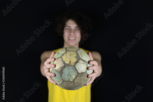 Smiling teen in a yellow shirt presents well-used soccer ball toward the camera on dark background, conveying sport, pride, teamwork. The teenage boy always plays football. Focus on the ball