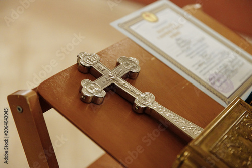 Fotografi Silver Orthodox crucifix resting on a wooden church stand next to a golden Gospel book and a religious certificate during a sacred ceremony