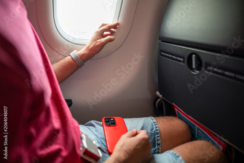Photos Male tourist passenger awaiting departure sitting in airplane beside window with opened blinds, man traveler with smartphone looking outside porthole waiting while plane getting ready for flight