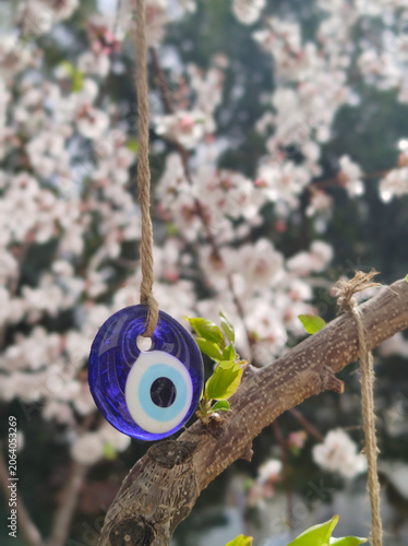A close-up vertical shot of a classic glass evil eye charm (nazar boncugu) hanging from a tree branch, featuring a beautiful soft-focused background of blooming white spring flowers.