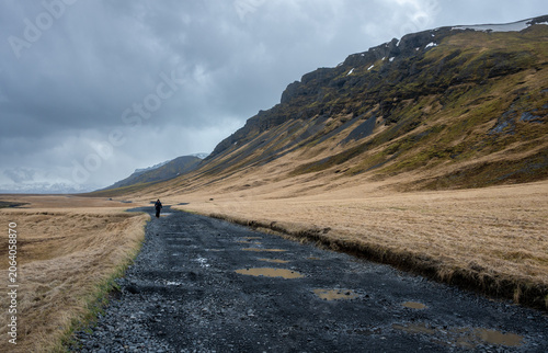 Fotografija Hiker trekking along a desolate gravel f road in a vast valley, surrounded by mountains with patches of snow and dry grass