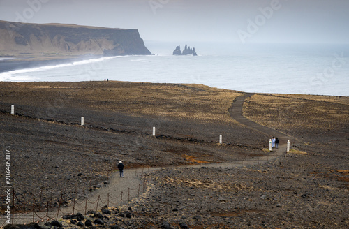 Hikers follow a winding path across the dyrholaey peninsula toward reynisfjara black sand beach with reynisdrangar sea stacks. Iceland landscape
