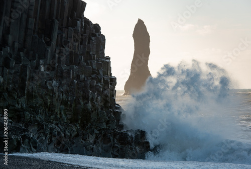 Reynisfjara beach showing powerful ocean waves crashing onto dark basalt columns with a distant sea stack under a bright sky. Vik Iceland