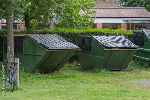 Two large green dumpsters sit in a grassy area with a building in the background.