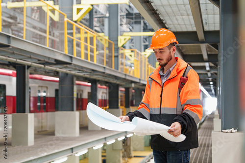 Construction engineer intently examines architectural plans on bustling subway platform during vibrant building construction project; he wears hard hat and orange safety vest for site safety.