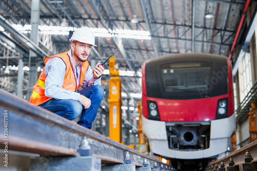 Industrial railway technician performs detailed inspection of electric train within expansive workshop utilizing handheld radio and standard safety apparel – white helmet, orange vest, light-colored s