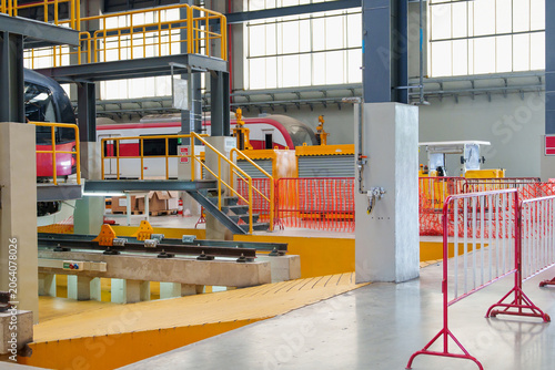 Detailed inspection of red electric train underway within large industrial garage facility featuring extensive machinery and prominent orange safety barriers for thorough maintenance work.